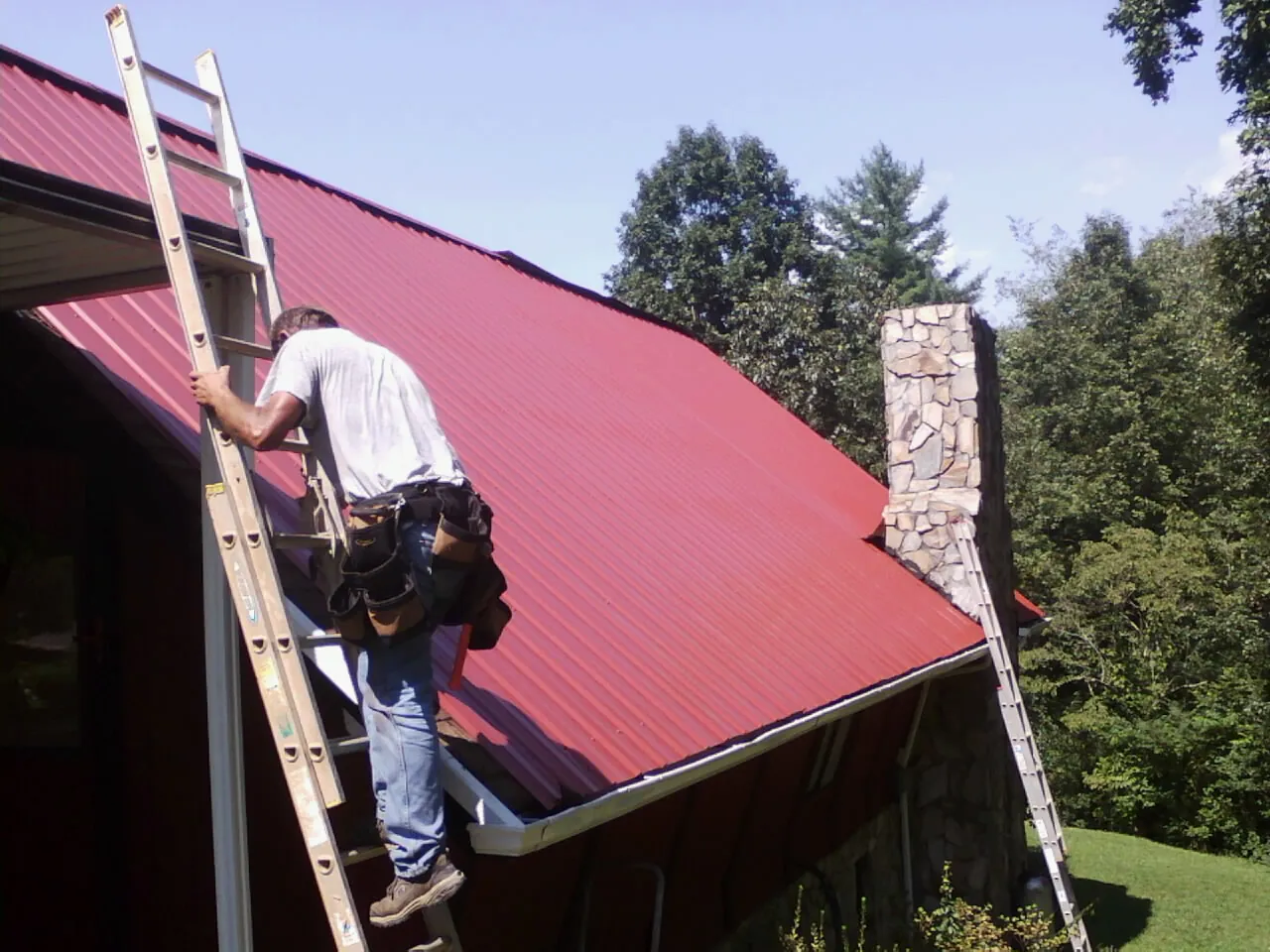 Roofer on a ladder beside a red metal roof and stone chimney — Soffit & Fascia Repair by Roofing Brothers in Cripple Creek
